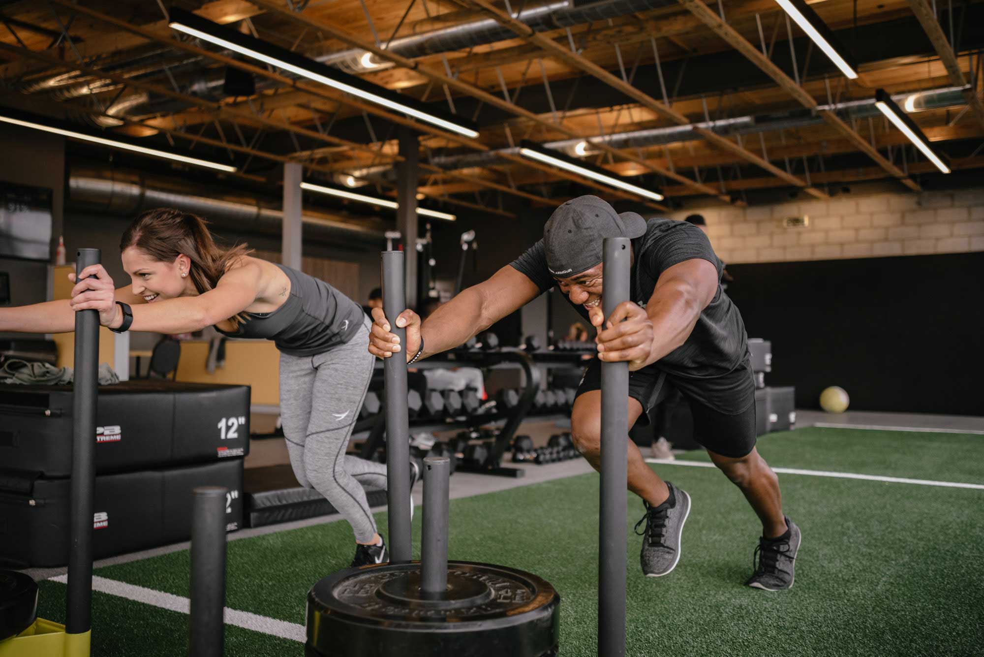 Two CF/THRIVE members pushing weighted sleds side by side on the turf at City Fitness